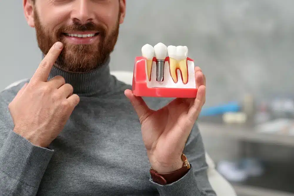 Smiling patient holding a dental implant model that illustrates how implants replace missing tooth roots and support long-term jawbone health.